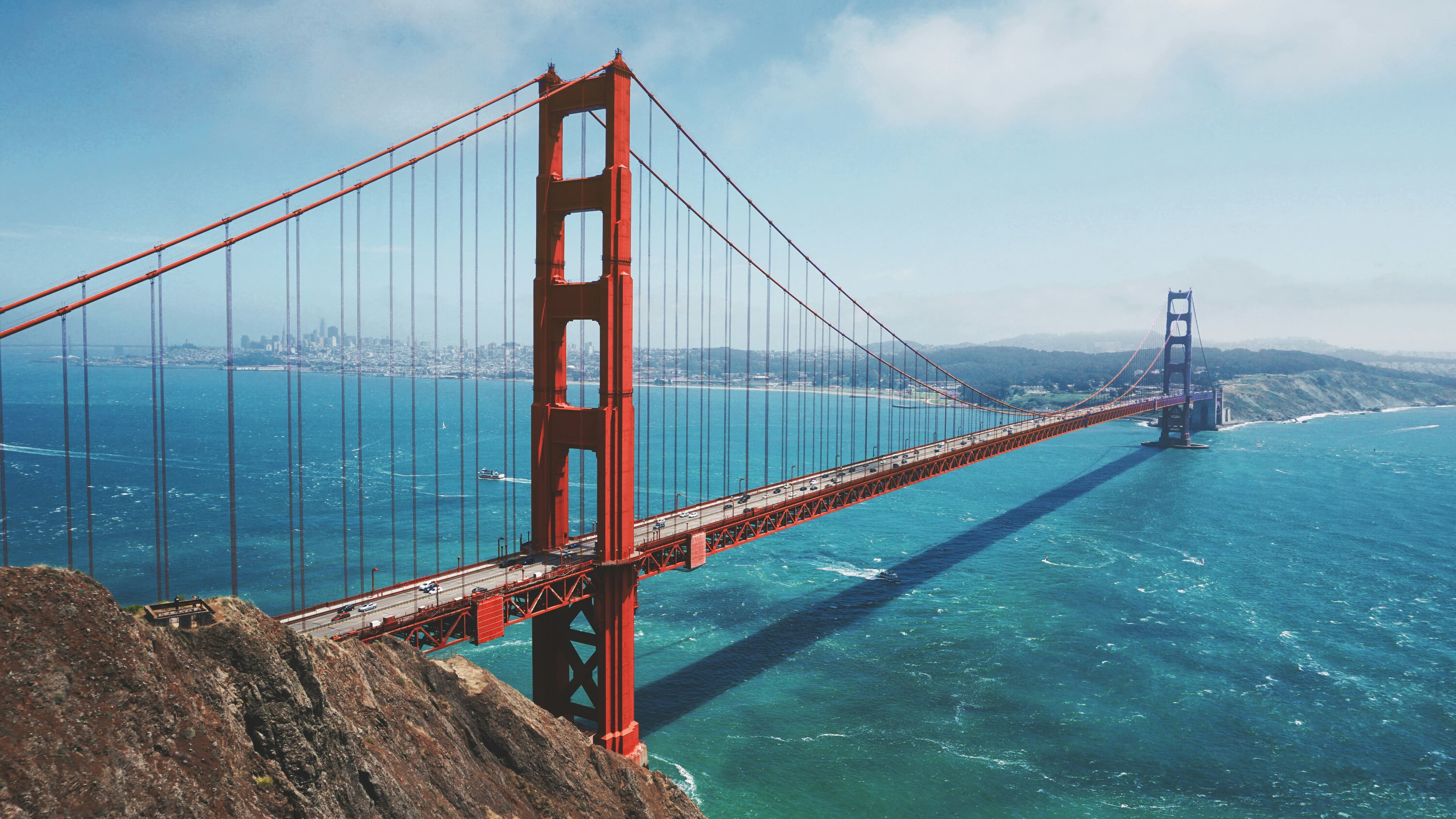 Golden Gate Bridge spanning blue water on a bright day, seen from a rocky bluff