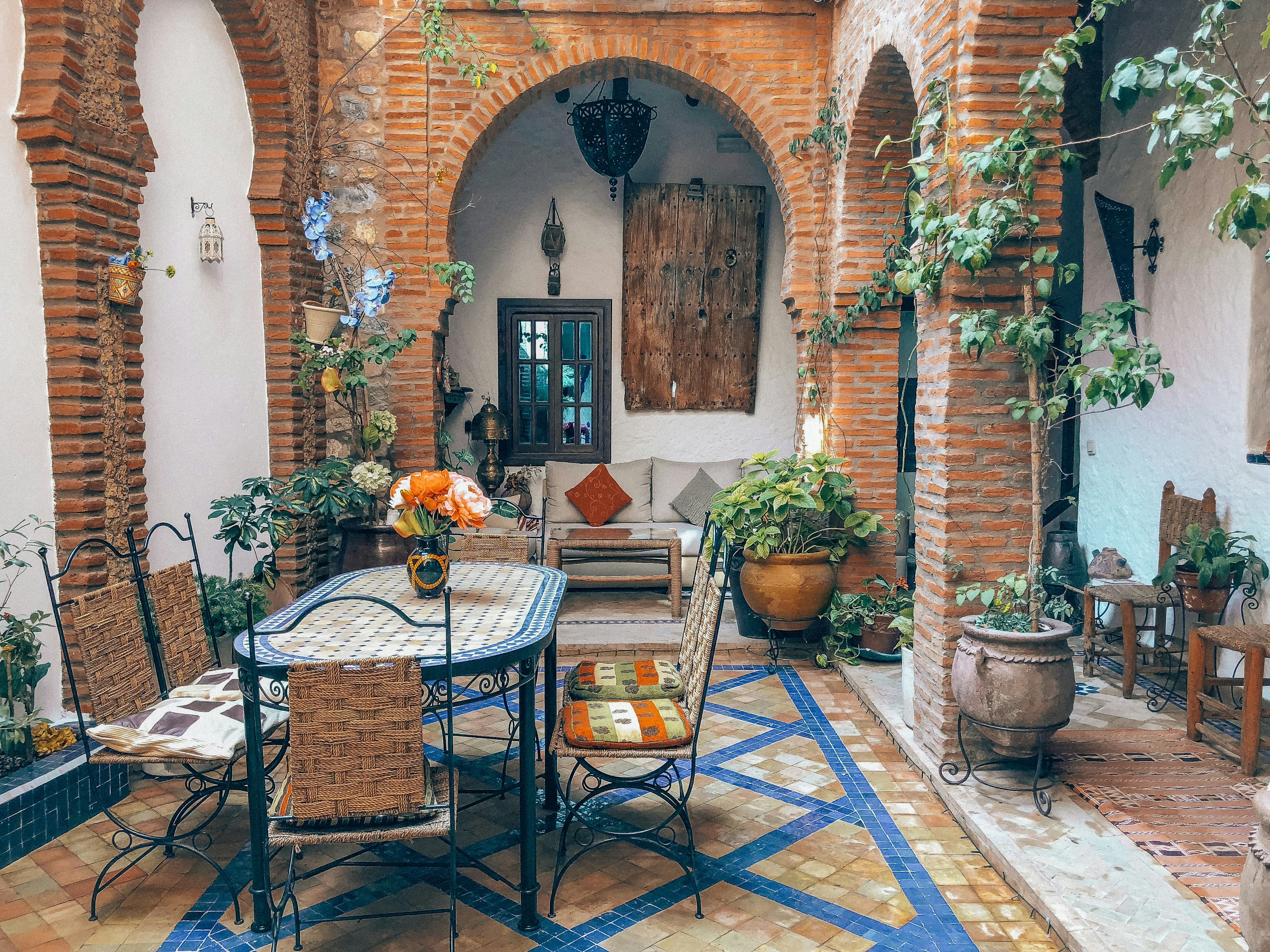 Patio d'un riad marocain : arcades de brique, zellige bleu et blanc, table en mosaïque et plantes en pot