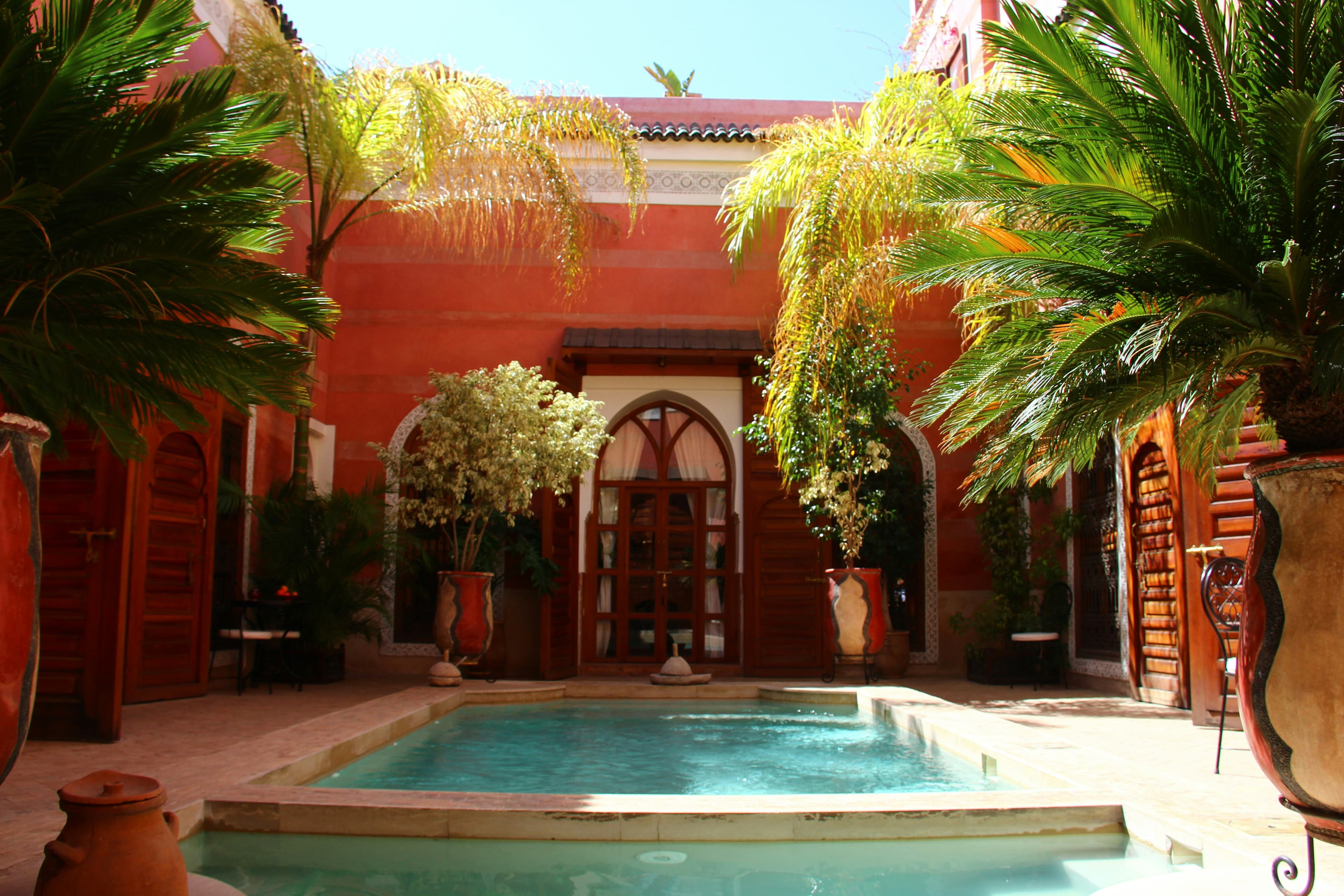 Riad aux murs rouges autour d'une piscine bordée de palmiers, sous un ciel clair