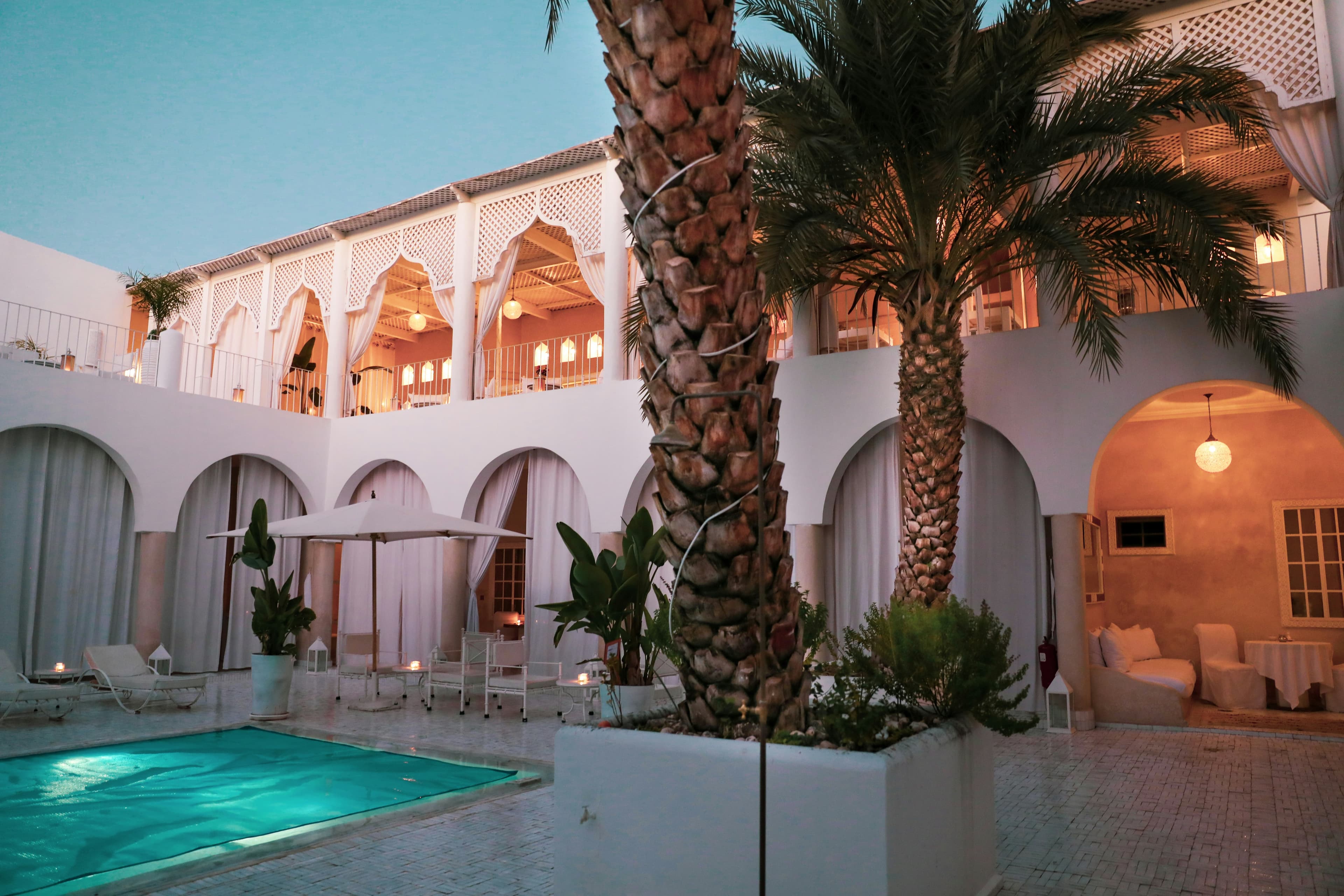 Patio d'un riad blanc au crépuscule, bassin turquoise, deux palmiers et arcades éclairées