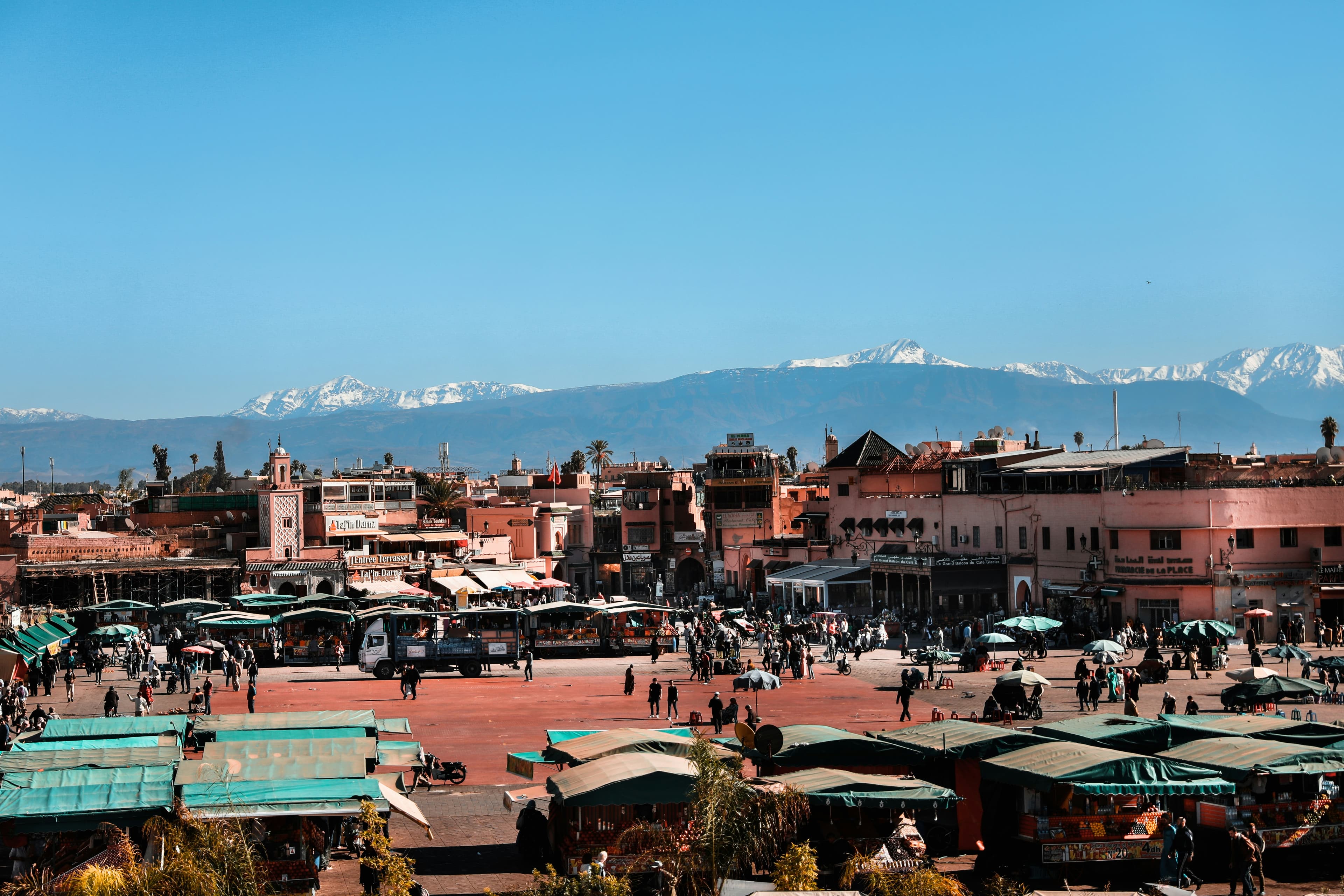 La place Jemaa el-Fna à Marrakech, ses étals sous auvents et ses toits ocre, avec l'Atlas enneigé à l'horizon